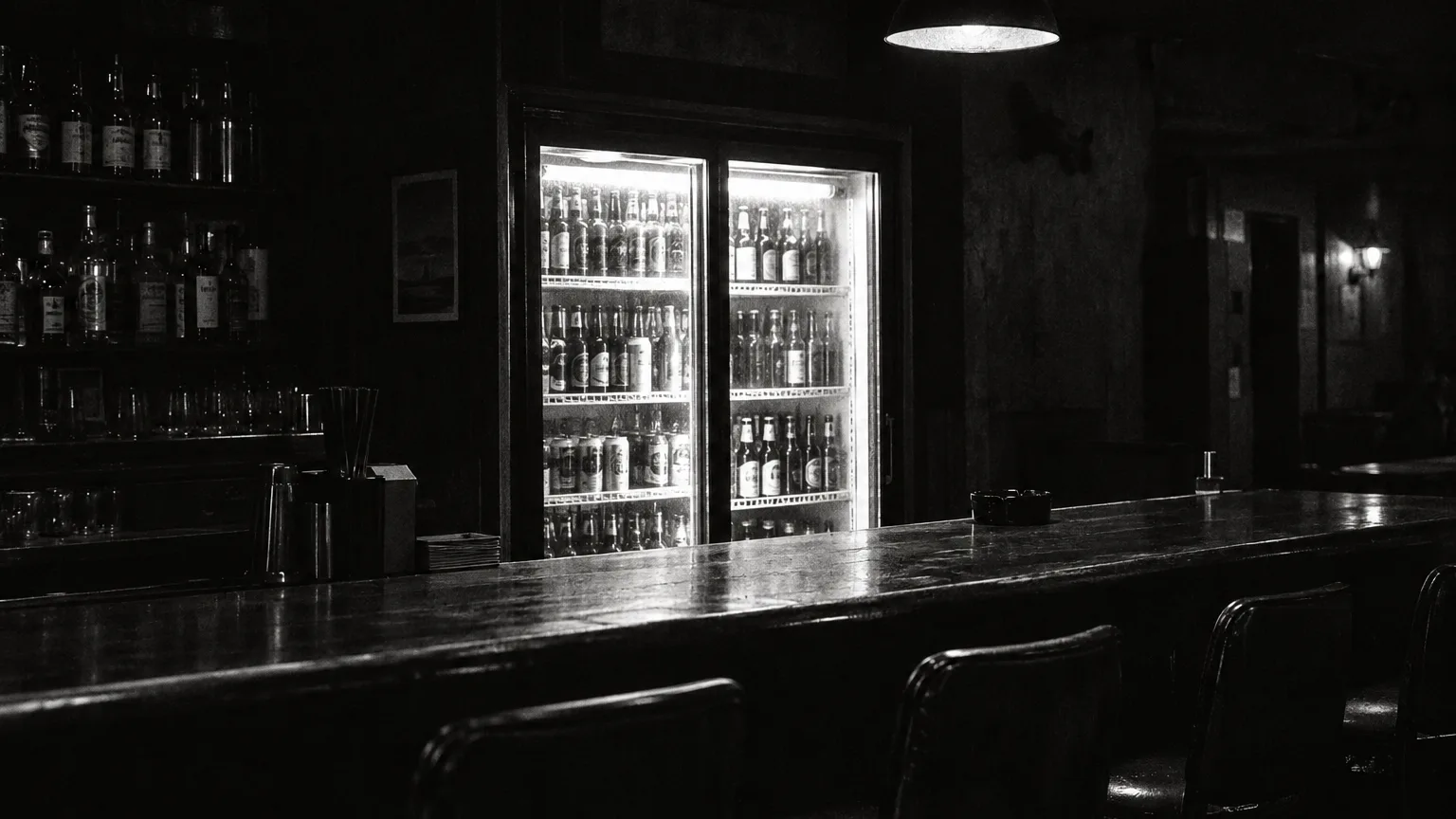 Black and white cinematic image of a glass door bottle cooler humming behind an empty dive bar counter
