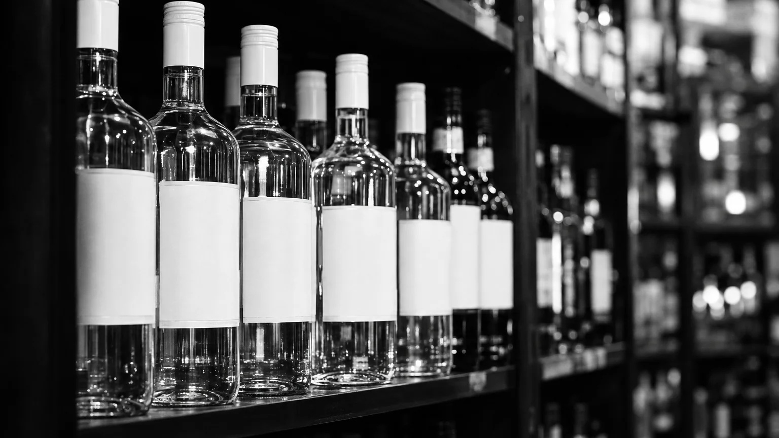 Black and white image of clear rum bottles on a shelf, white labels bright against dark shelving