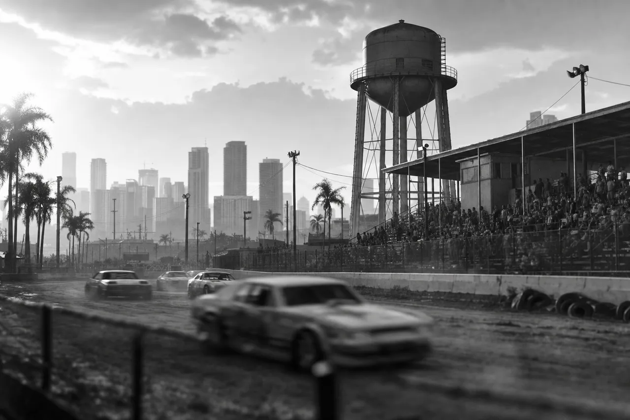 Black and white cinematic view of a rural speedway with a water tower, weathered grandstands and blurred stock cars