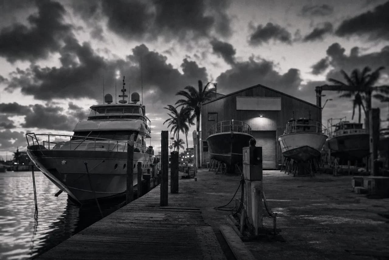 Black and white Florida Keys boatyard at dusk with a yacht moored at a wooden dock and boat hulls up on dry-dock cradles