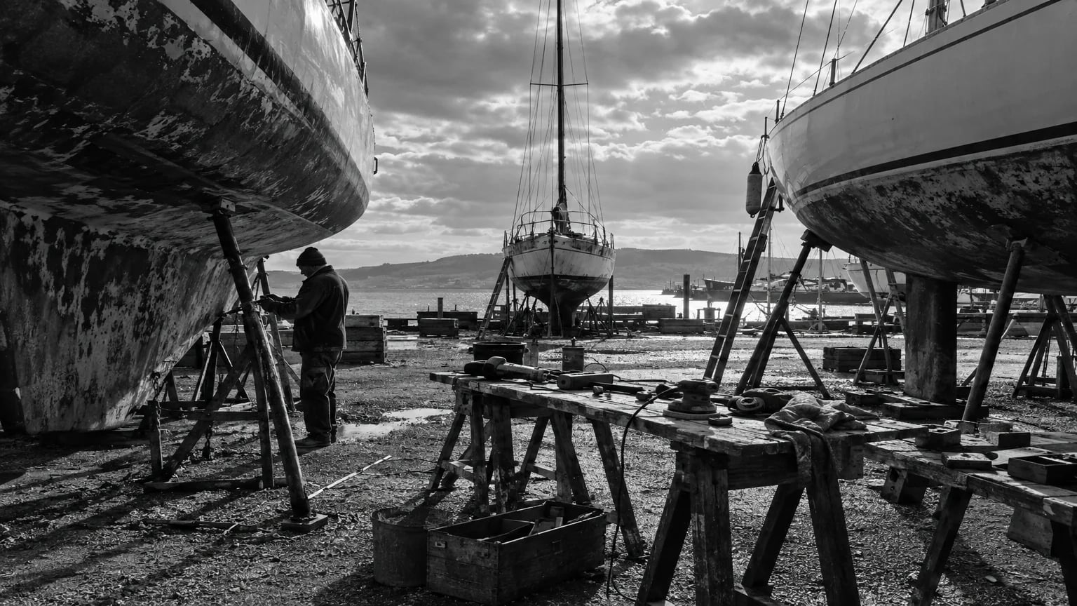 Black and white cinematic photograph of a boatyard with hulls up on stands and tools scattered on trestles