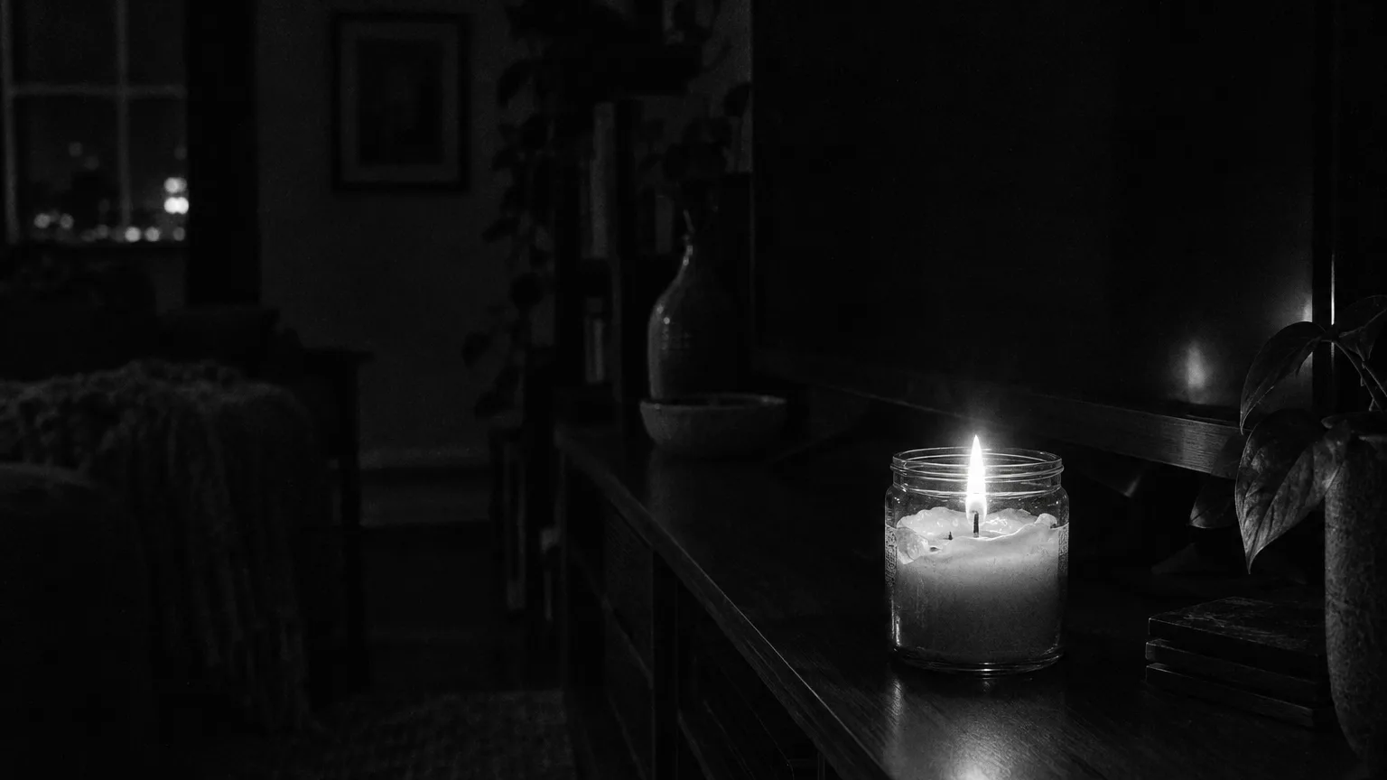 Black and white cinematic close-up of a lit jar candle on a TV stand, wax pooling around the wick