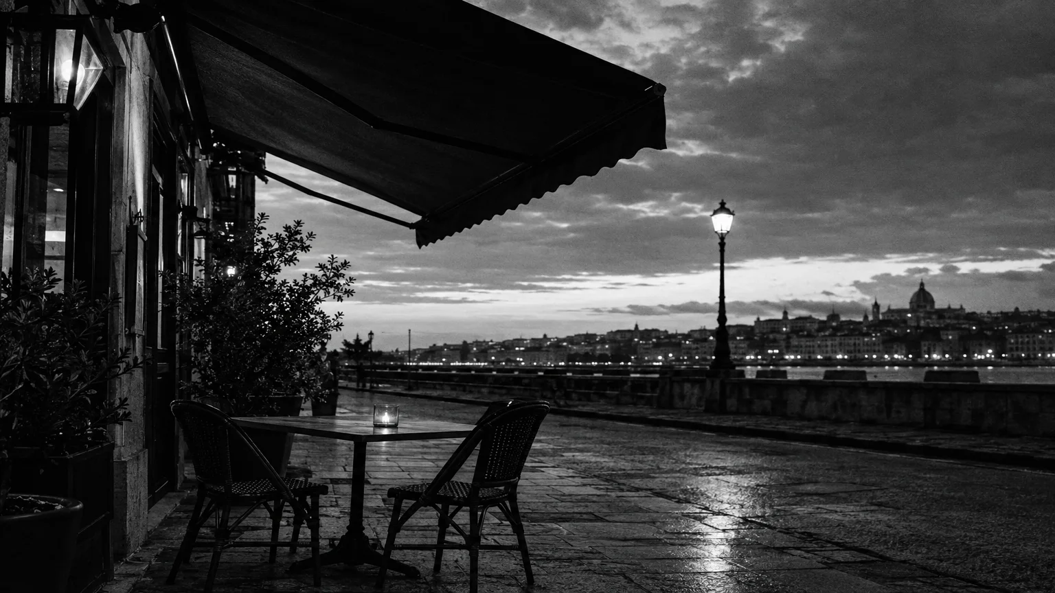 Black and white cinematic photograph of a trattoria awning over an empty pavement table in evening light