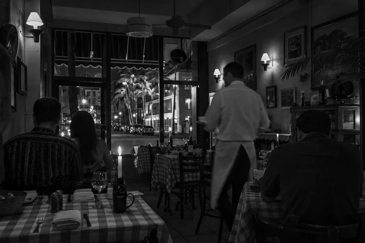 Black and white cinematic view of an Italian restaurant with checked tablecloths, a candle in a wine bottle and plates of pasta