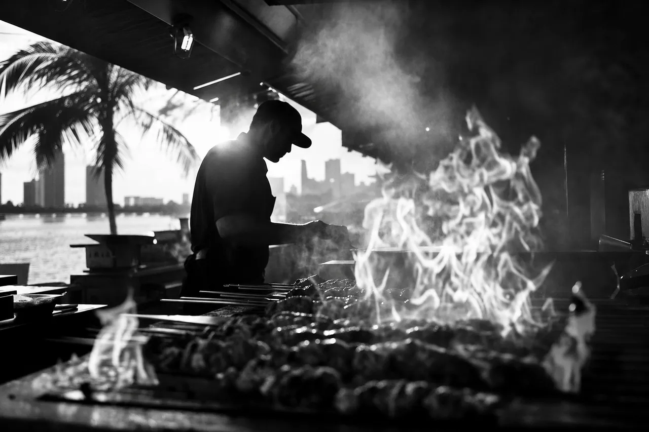 Black and white cinematic shot of flames leaping around skewers on an open grill with a chef silhouette in smoke