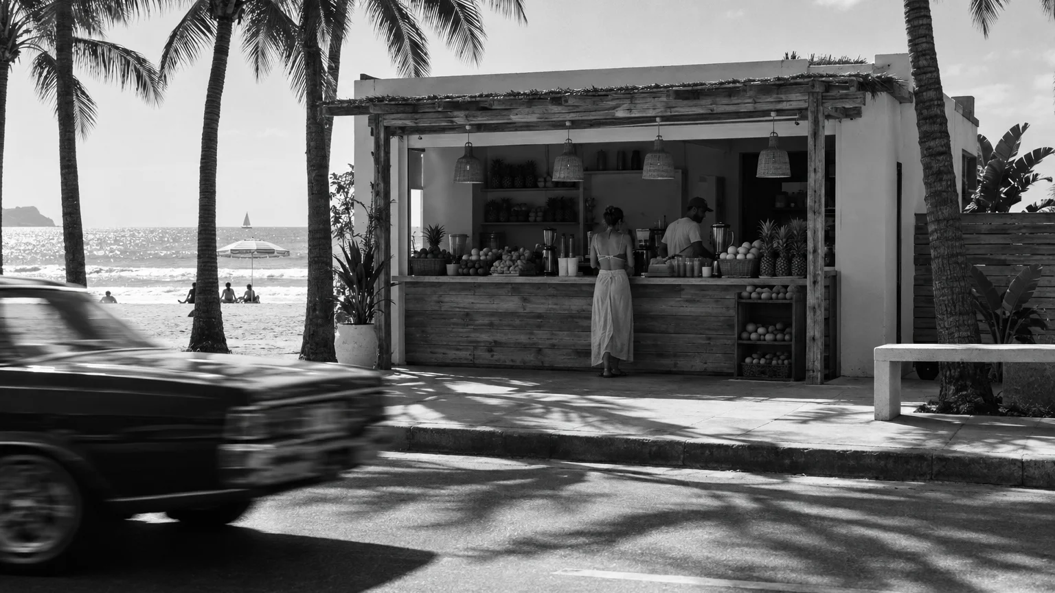 Black and white photo of a small juice bar storefront on a beach strip, blurred by a passing car