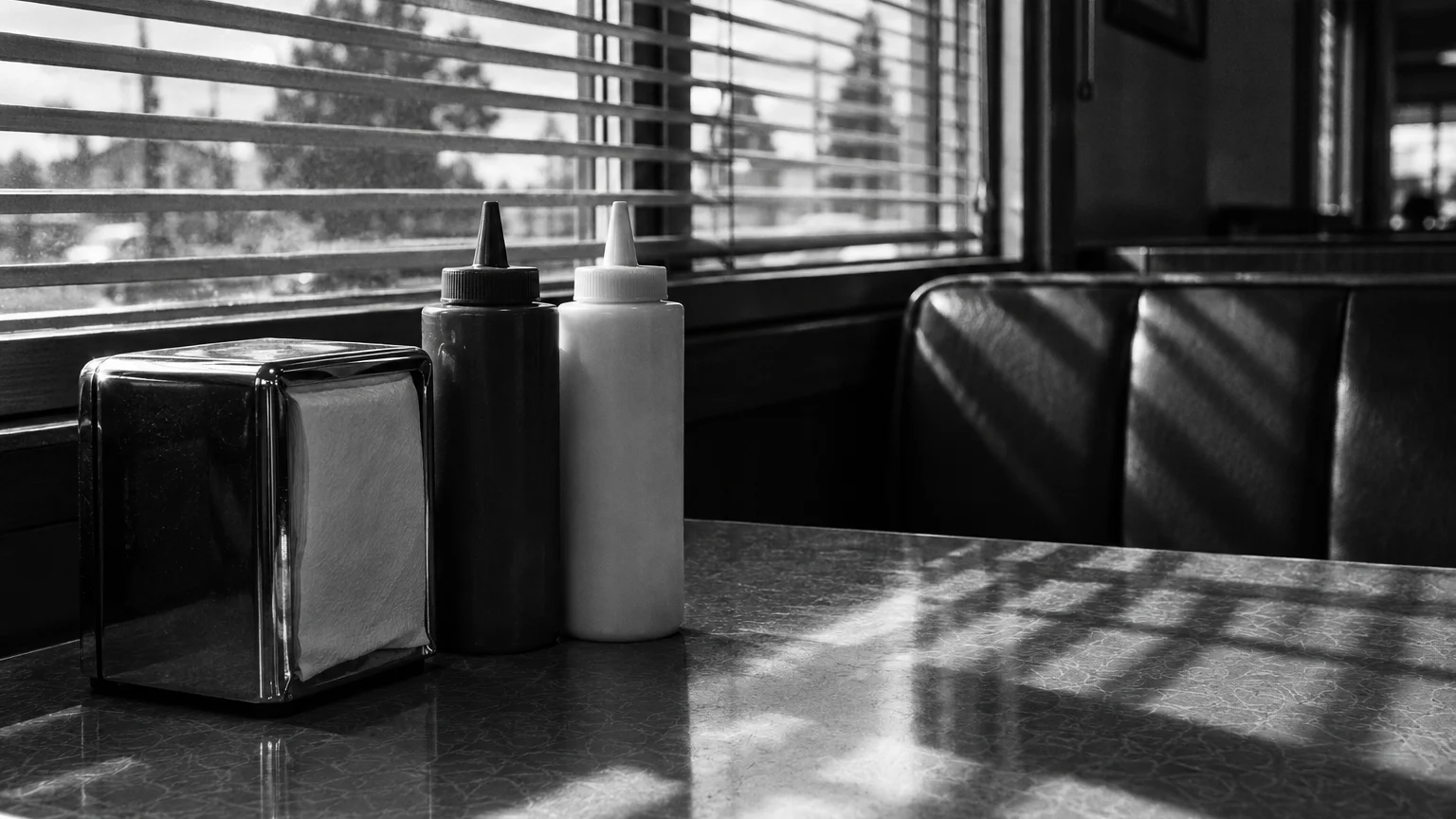 Black and white cinematic photograph of ketchup and mayonnaise squeeze bottles on a diner table by a window