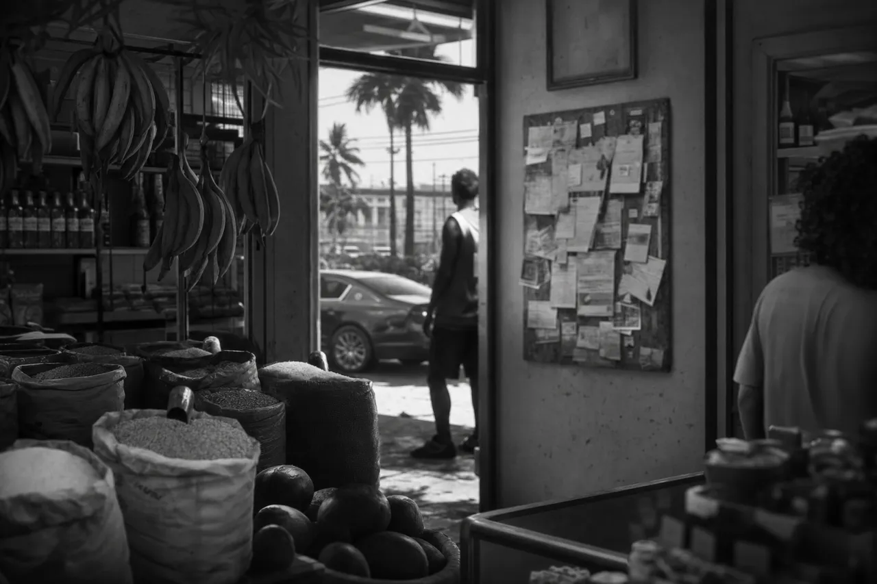 Black and white cinematic view of a Caribbean grocery with spice sacks, hanging plantains and a flyer-covered notice board