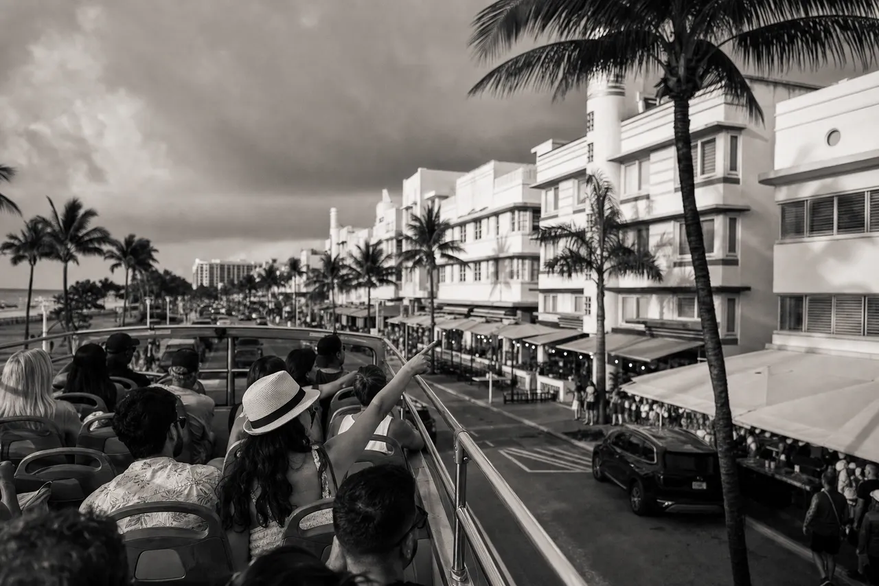 Black and white cinematic view of an open-top sightseeing bus on an ocean drive with tourists pointing from the upper deck