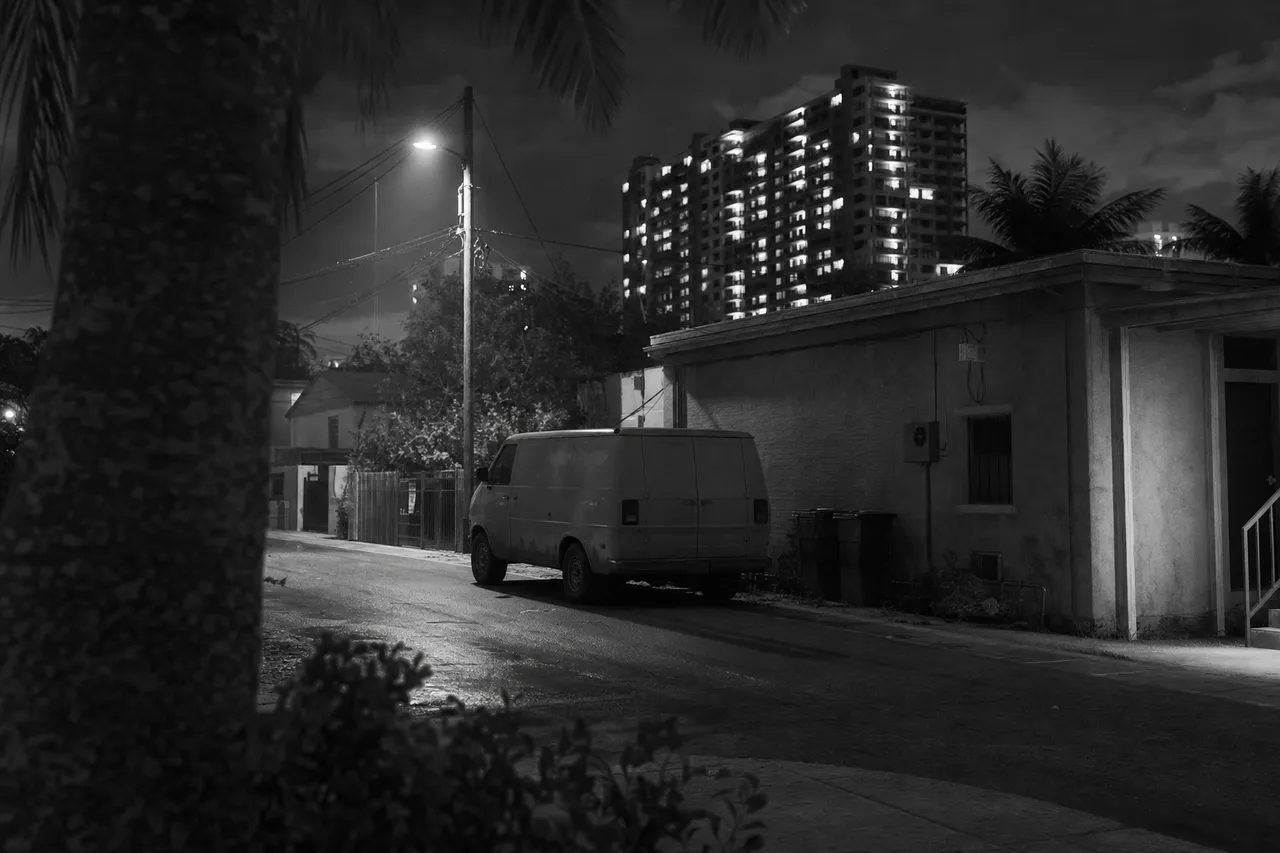 Black and white cinematic night view of an anonymous panel van in a narrow side street under a single streetlamp