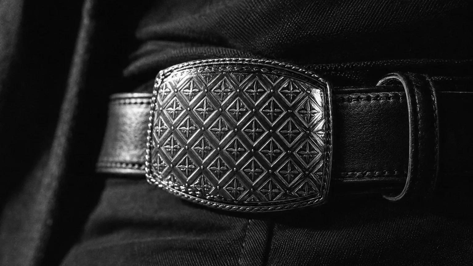 Black and white cinematic close-up of a monogram patterned belt buckle catching studio light