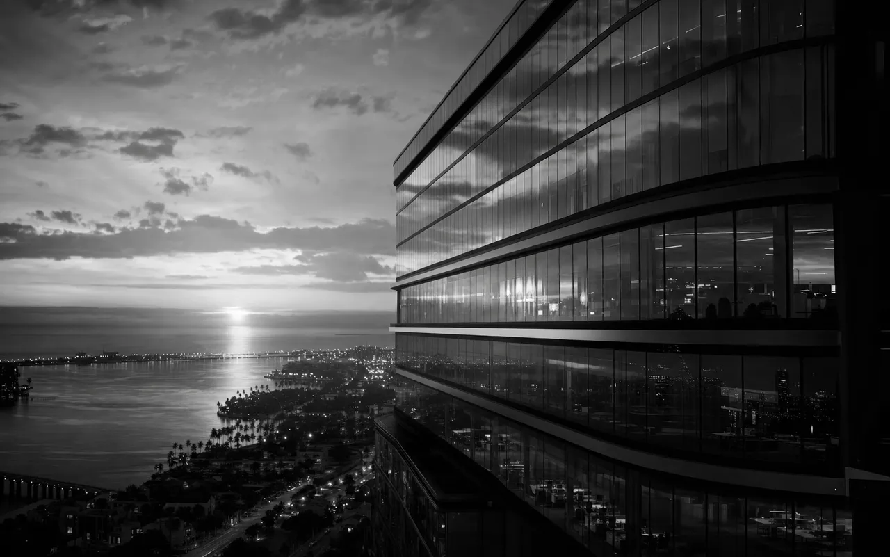 Black and white cinematic view of a waterfront skyscraper with a sunset reflected across its glass curtain wall