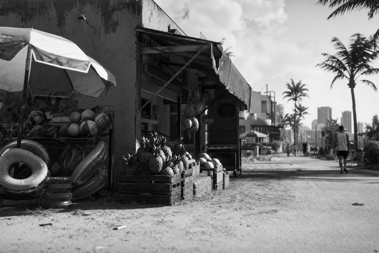 Black and white cinematic view of a small beach-block market with fruit crates and beach toys out front
