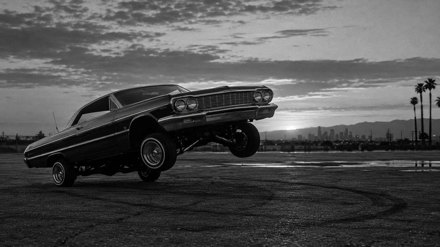 Black and white cinematic photograph of a lowrider car raised on one side in an empty lot at dusk