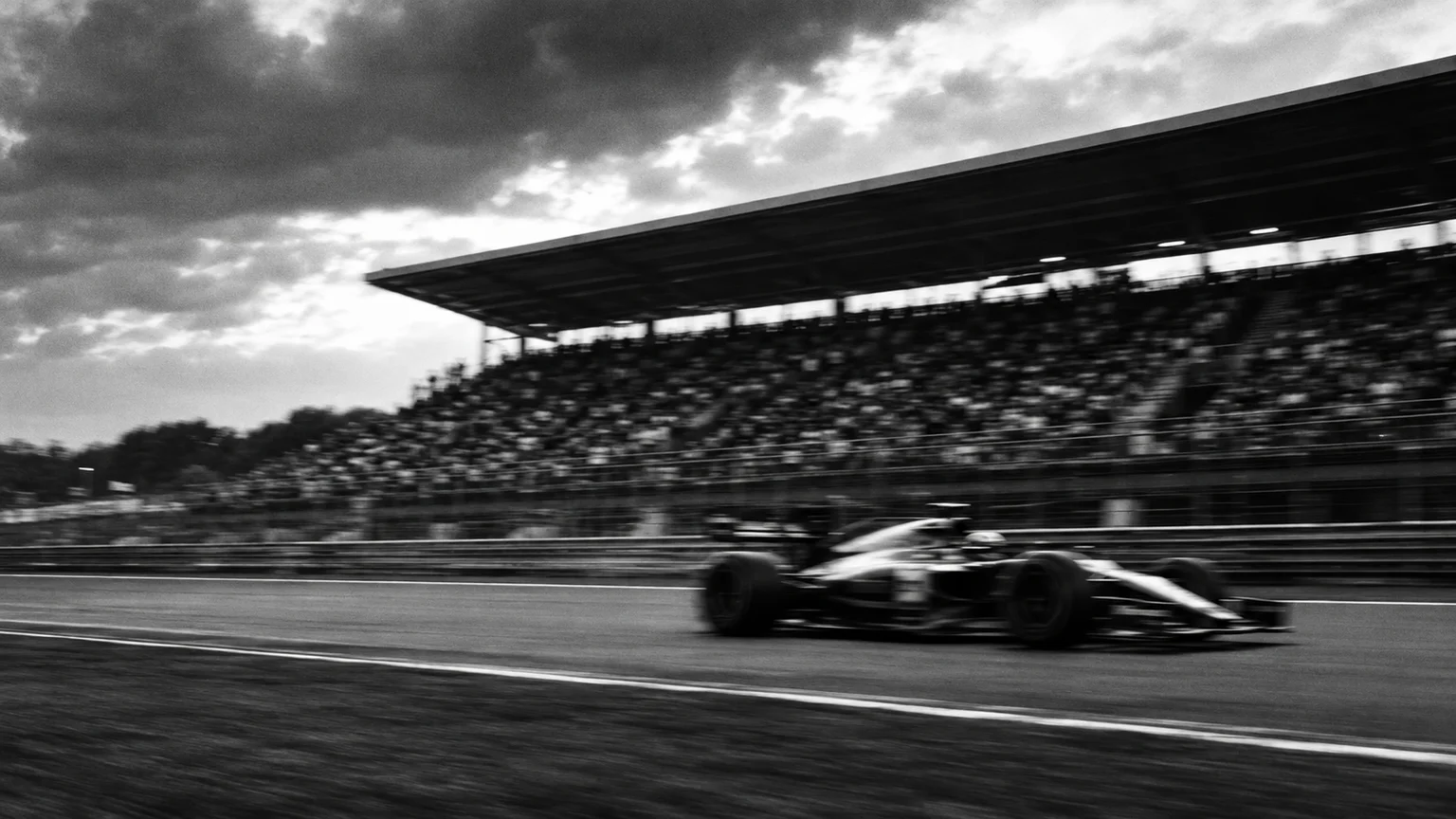 Black and white cinematic photograph of a blurred race car passing a grandstand, sponsor boards unreadable