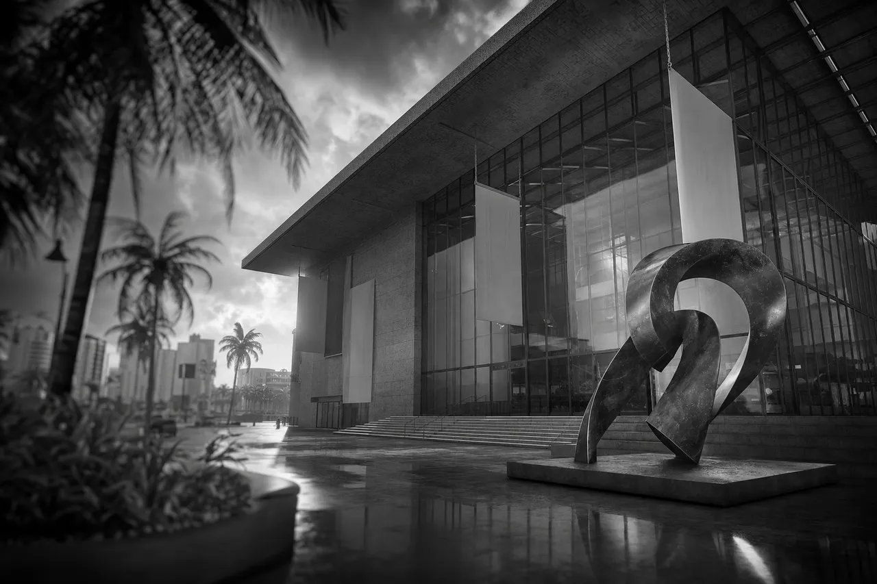 Black and white cinematic view of a modern art museum facade with blank banners and an abstract plaza sculpture