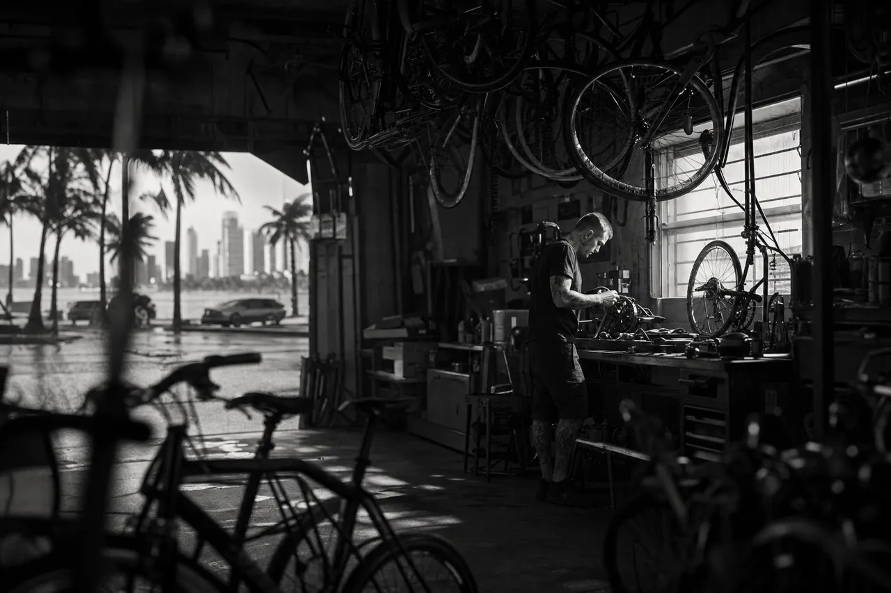 Black and white cinematic view of a bicycle shop with bikes hanging from hooks and a mechanic at a workbench
