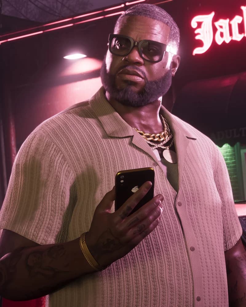 Boobie Ike standing outside the Jack of Hearts adult entertainment club at night, wearing a cream knit shirt and a heavy gold chain, holding a phone.