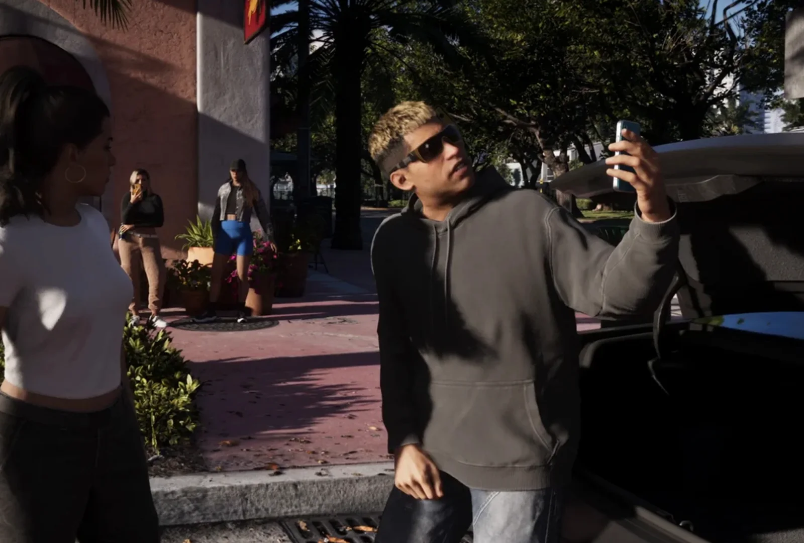 Camillo filming himself on his phone beside the open trunk of a car on a Vice City street, blond hair and yellow-framed sunglasses, grey hoodie.