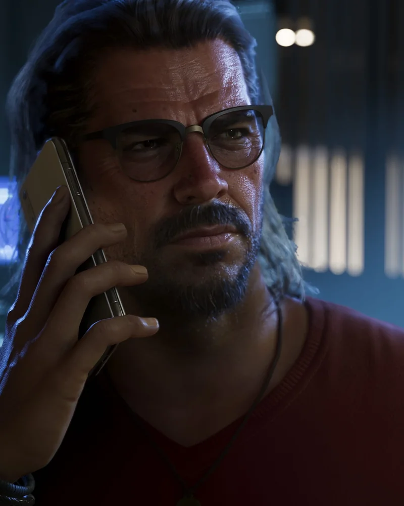 Raul Bautista in close-up, wearing thin-rimmed glasses and a red v-neck, holding a gold phone to his ear inside a dim control room.