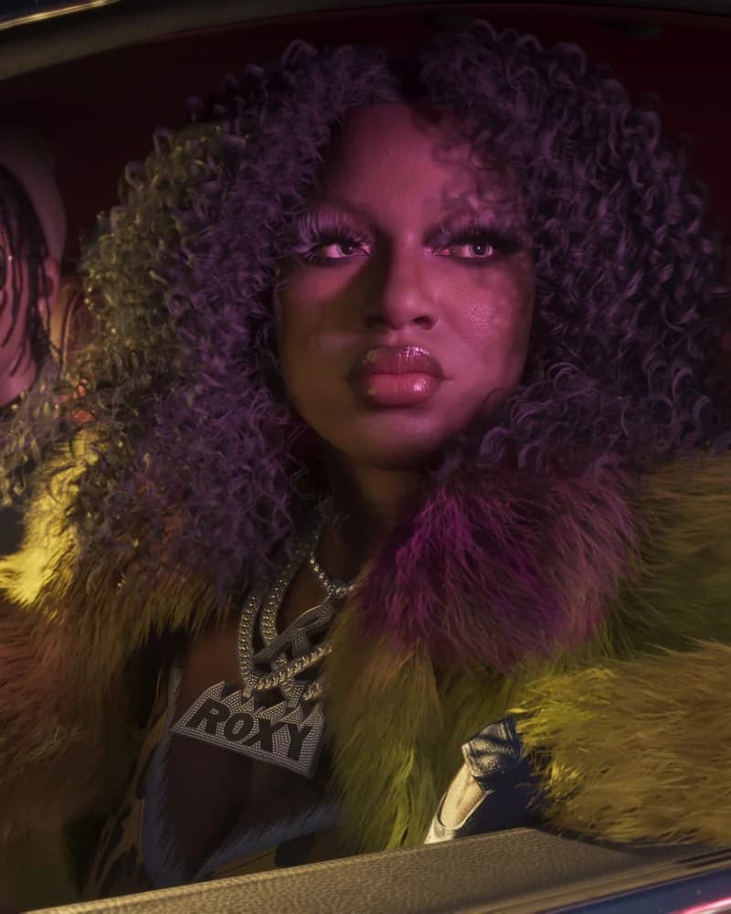 Roxy in the passenger seat of a car at night, wearing a yellow and purple fur coat with a chunky "ROXY" diamond necklace, hair lit purple by neon.