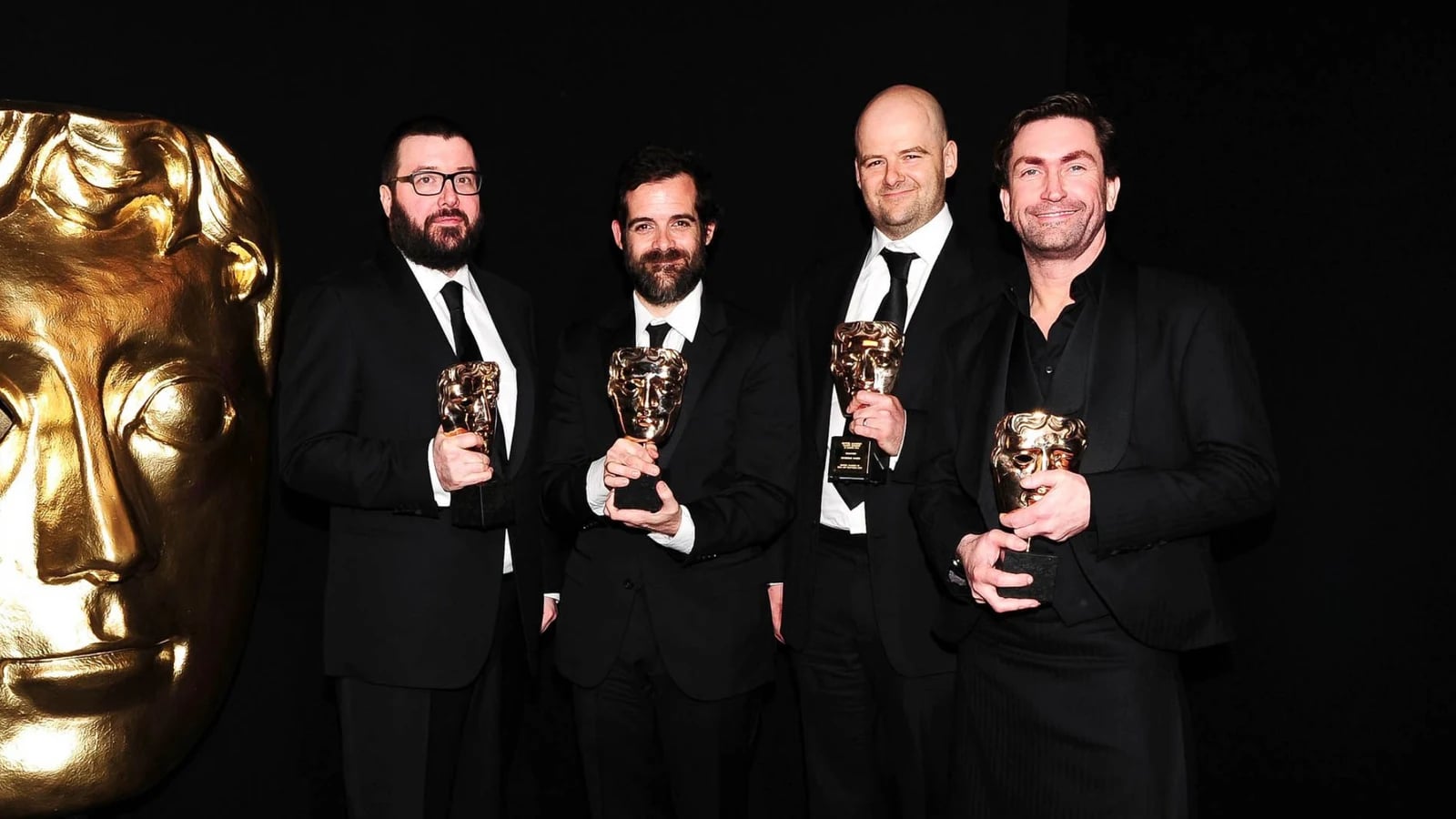 Rockstar Games leadership including Dan Houser and Leslie Benzies holding BAFTA awards beside a giant golden BAFTA mask