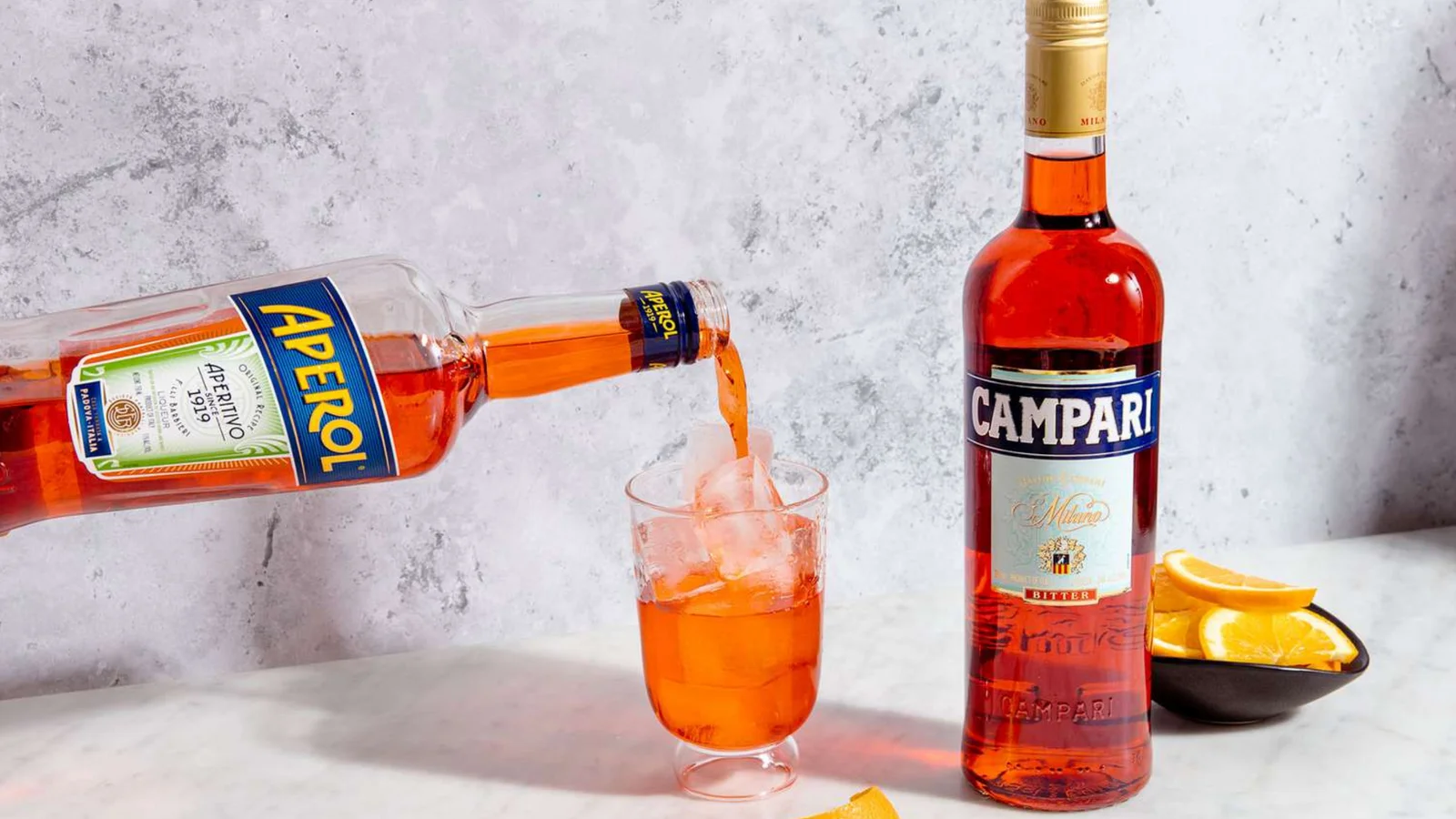 Aperol being poured into a glass next to a Campari bottle