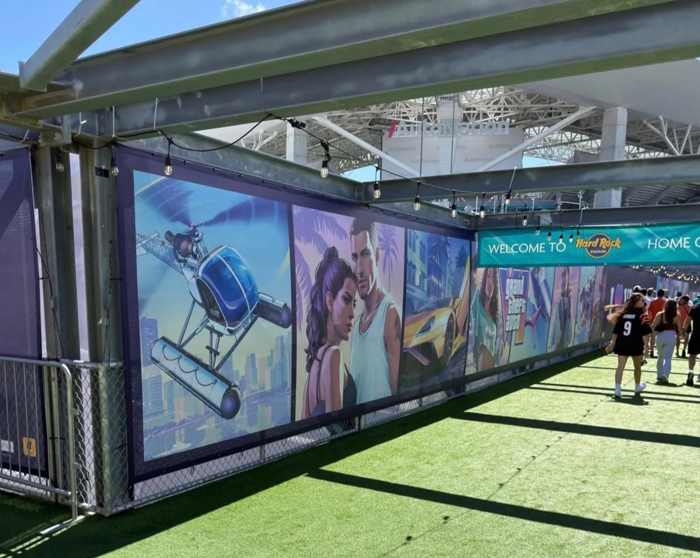 GTA 6 cover art wrap at Hard Rock Stadium showing the helicopter, Jason and Lucia and the yellow supercar along a fence line