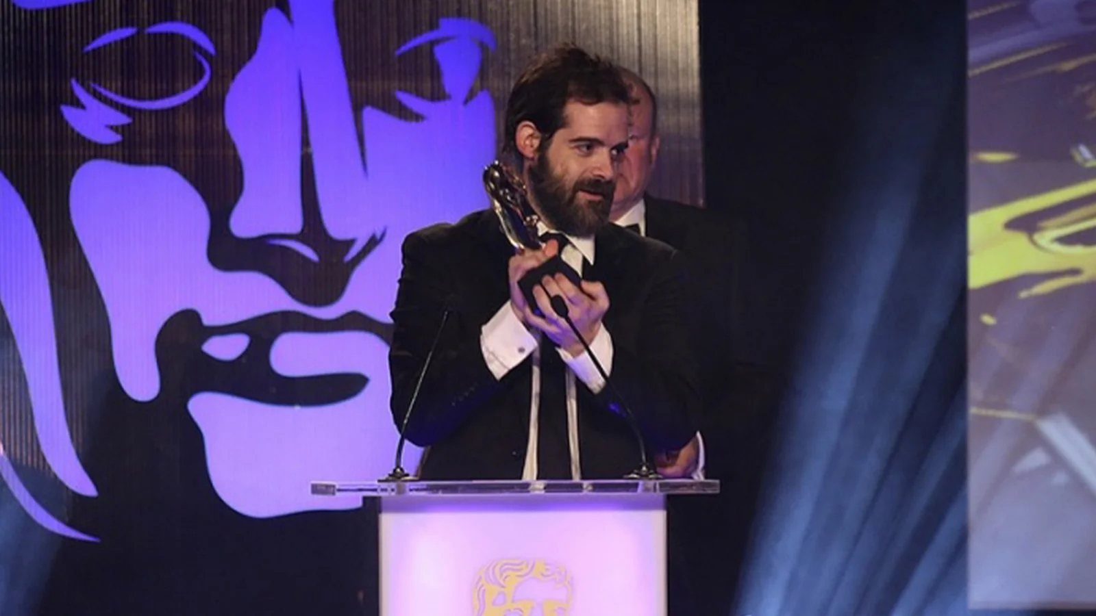 Sam Houser accepting a BAFTA award at a ceremony podium in front of a giant BAFTA mask backdrop